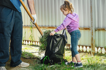 adults and children do household chores together, remove garbage, leaves in the yard of the house. Father collects garbage with a shovel, a little girl helps him and holds a garbage bag