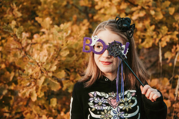 Fototapeta na wymiar Portrait of cute smiling caucasian teen girl with loose hair wearing black hoodie, violet boo glasses and headband with black spider. She is hiding one eye with magic wand. Halloween decoration.