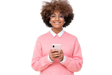 Smiling pretty african girl with afro hairstyle holding smartphone with both hands, looking at...