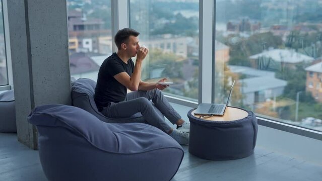 Young self-employed man having lunch break resting from work at laptop. Male drinking coffee and enjoying city view from a big window.