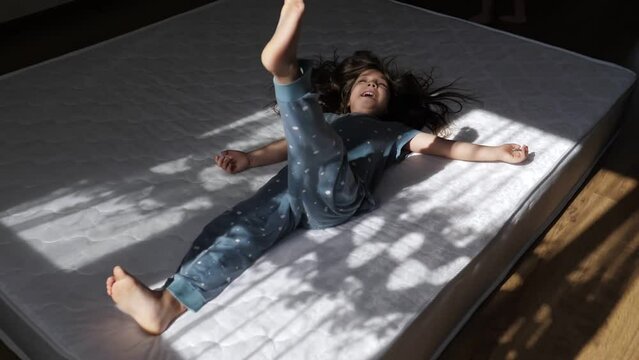 Happy 5 year old girl in pyjamas falling on the mattress with shade from the curtain. Kid spending active energetic funny time in the morning in bedroom. Happy childhood, concept of new cozy home.