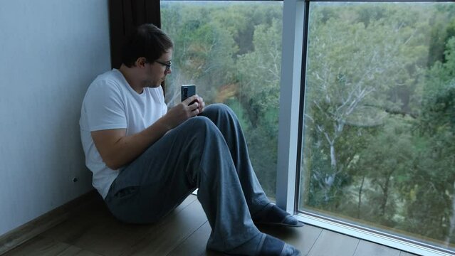 Sad young man sitting on the floor looking through the window.