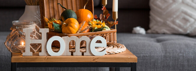 Cozy autumn concept. Home warmth in cold weather. Still-life. Pumpkins, wicker basket, candles, flowers and the inscription home on the coffee table in the home interior of the living room.