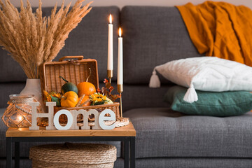 Fototapeta na wymiar Cozy autumn concept. Home warmth in cold weather. Still-life. Pumpkins, wicker basket, candles, flowers and the inscription home on the coffee table in the home interior of the living room.
