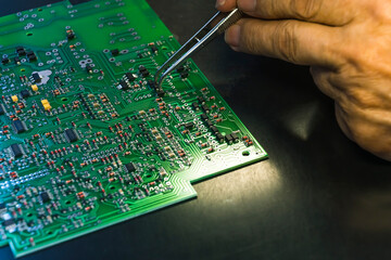 Hand gripping tweezers assembling electrical parts on detailed surface of printed circuit board...