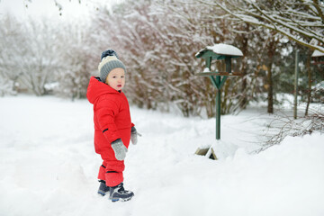 Fototapeta na wymiar Adorable toddler boy having fun in snow covered park on chilly winter day. Child exploring nature.