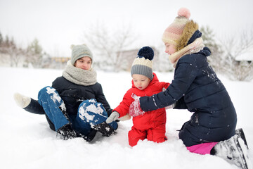 Two cute young sisters and their toddler brother having fun in snow covered park on chilly winter day. Children exploring nature.