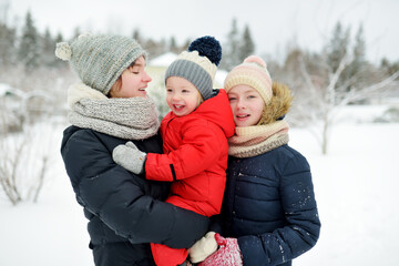 Two cute young sisters and their toddler brother having fun in snow covered park on chilly winter day. Children exploring nature.