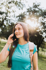 Teenage student girl wearing aquamarine t shirt, making a phone call to her family in the park. Sun backlight