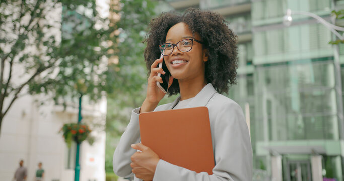 African woman with curly hair talks phone outdoors near business center, she hopda a laptop computer and a smartphone - Powered by Adobe