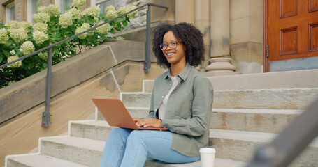 Curly Afro American young woman, seats on the stairs and working outdoors with her 13-inch personal laptop computer, she having her coffee near by
