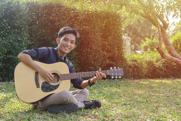 Portrait of attractive Asian man in casual shirt playing guitar sitting on garden grass