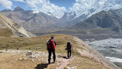 Fototapeta premium Two tourists with big backpacks are walking in the mountains. View of the rocky mountains, snow-capped peaks and hills of Kyrgyzstan.