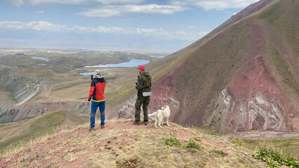 Fototapeta premium Two tourists with a dog stand on a high hill, look into the distance and take a photo on the phone. View from a height of colorful rocky mountains, lakes, green slopes and hills of Kyrgyzstan. 
