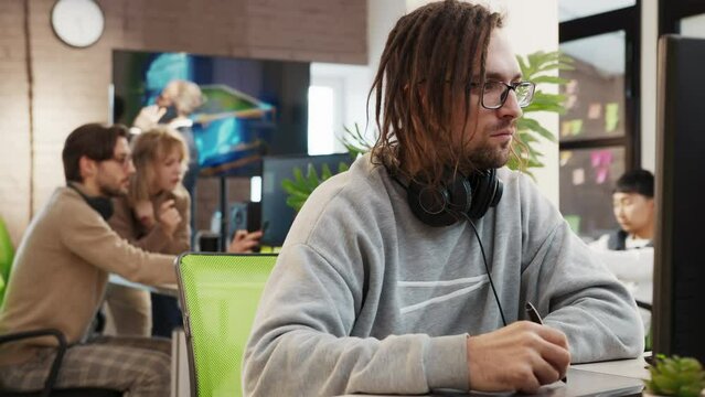 Caucasian male artist designer with dreadlocks and glasses draws on a tablet a 3D model for a computer game in the office of a project development studio. 
