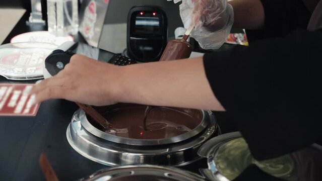 Close-up of orange fruity ice cream dipped in liquid chocolate glaze, female worker making and selling delicious glazed ice-cream on stick in food truck, sales stand outdoors, city park street shop.