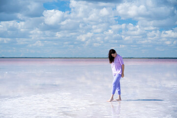 Fototapeta na wymiar Bursol pink lake. A woman in lilac clothes walks along the bottom of the salt of the lake. Clouds are reflected in pink water. Concept: freedom, looking into the bright future, lightness, enjoying the