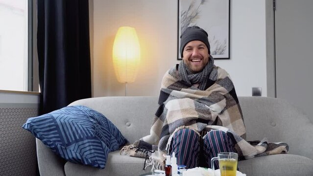 Funny cheerful young man in a hat suffering from a common cold and flu or allergy sit at home wrapped in blanket. Healthcare concept