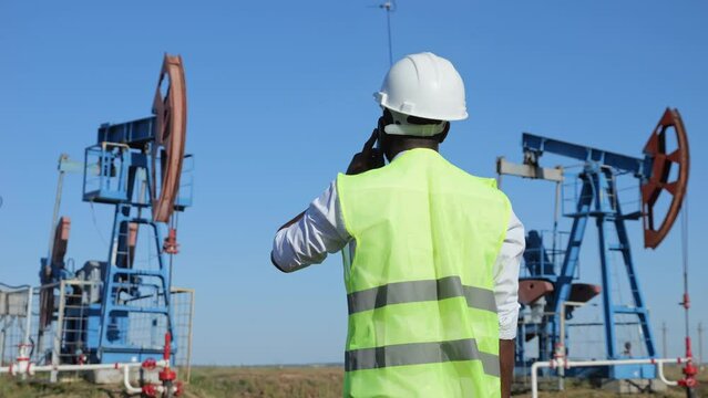 African American man talks on phone about beam unit operation controlling production of crude oil. Black engineer makes report about working jack-pump
