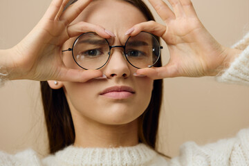 an attractive woman stands on a light brown background in a white knitted sweater and black glasses on her face, leaning her hands against her head, as if examining something, puffing out her cheeks