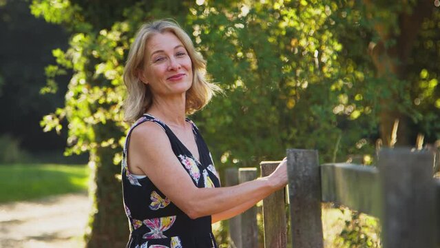 Casually dressed smiling mature woman leaning on wooden fence on walk in summer countryside - shot in slow motion