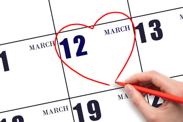 A woman's hand drawing a red heart shape on the calendar date of 12 March. Heart as a symbol of...