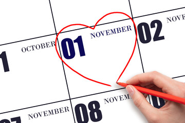 A woman's hand drawing a red heart shape on the calendar date of 1 November. Heart as a symbol of...