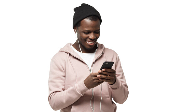 Young handsome black guy in pink hoodie and black hat, listening music with headphones and looking at screen of his smartphone