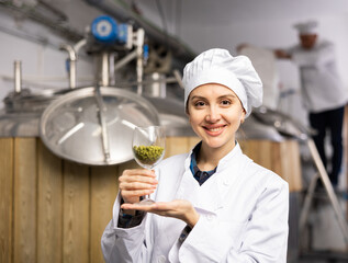 Portrait of positive woman brewmaster holding glass of hops pellets in beer factory.