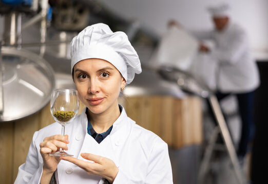 Portrait of positive woman brewmaster holding glass of hops pellets in beer factory. - Powered by Adobe