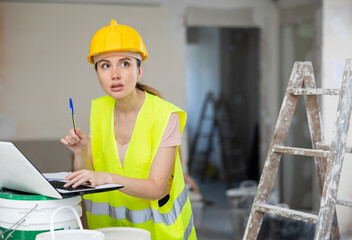 Female foreman in a protective helmet and a yellow vest checks the execution of repair work using a laptop.