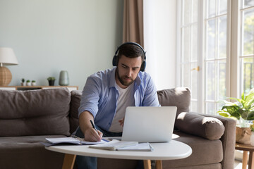 Concentrated millennial adult student man in wireless headphones studying online at home, watching,...