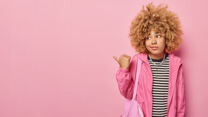 Horizontal shot of curly haired woman points thumb away on left shows promo offer looks with disapproval wears striped jumper and windbreaker isolated over pink background. Oh no look there.