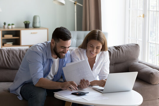 Cheerful young millennial husband and wife doing domestic paperwork, accounting job, reviewing paper bills, receipts at laptop computer, using calculator, paying mortgage, rent fees on internet