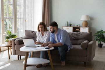 Serious millennial husband and wife discussing family expenses, using online payment laptop,...
