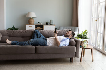 Cheerful millennial mobile phone user man chatting online, reading good news on screen, smiling, laughing, relaxing on sofa, lying on couch with smartphone at home, shopping on Internet. Full length