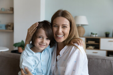 Cheerful loving mother and little son kid hugging on couch, looking at camera, smiling. Mother embracing cute preschool child with care, affection. Motherhood concept. Family head shot portrait