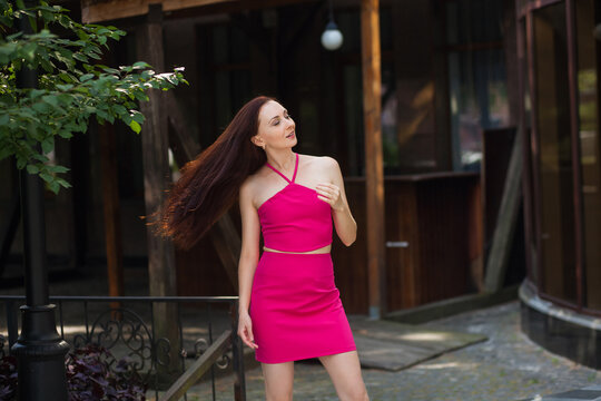 A stylish slender smiling woman with long dark hair, in a pink short sexy skirt and a tank top, stands near a modern building on a city street.