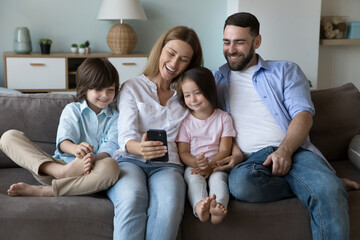 Happy family taking selfie on home couch. Cheerful millennial parents and two little children using mobile phone for video call, laughing, relaxing with gadget on sofa. Communication concept