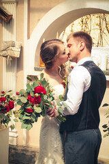 Stylish newlyweds at the wedding ceremony stand against the backdrop of a beautiful interior and kiss. The bride, in a light long dress, stands with a chic large wedding bouquet.