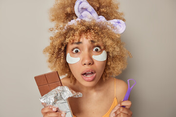 Headshot of amazed scared woman with bushy blonde hair holds tongue scraper and bar of chocolate stares with omg expression applies beauty patches to reduce wrinkles poses against grey background