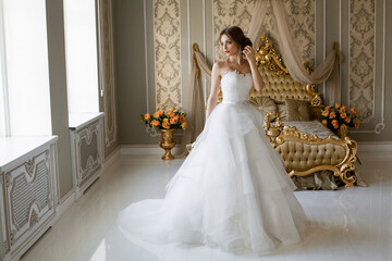 A beautiful smiling bride with chic makeup and hairstyle in a white wedding dress is standing in a bedroom with a stylish light vintage interior, in the morning before the wedding.