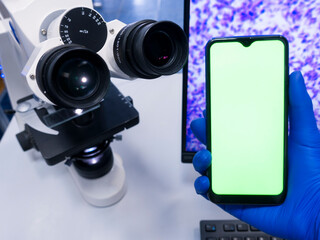 A gloved hand holds a smartphone with a green screen next to a large microscope. Transmission of images from a microscope to a phone. Connection to the microscope via smartphone.