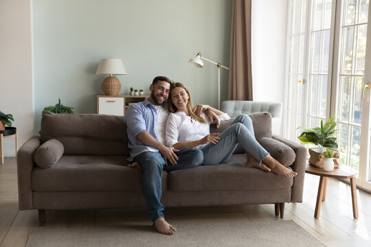 Happy young millennial couple of homeowners resting on comfortable sofa together, hugging, smiling, looking at camera, enjoying leisure. Cheerful husband and wife home full length portrait