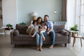 Happy millennial parents and two kids using mobile phone, resting on home couch together, holding...