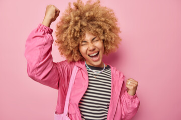 Overjoyed curly haired female model does winner gesture exclaims loudly celebrates success dressed in casual clothes feels ambitious and encouraged makes fist bump isolated over pink background