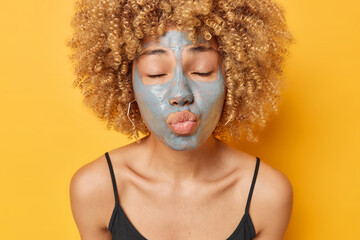 Funny curly haired woman stands with closed eyes keeps lips rounded applies beauty clay mask on face blows air kiss at camera dressed in black t shirt stands bare shoulders against yellow background