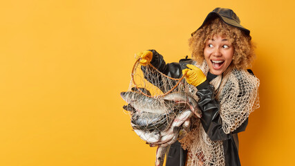 Successful fisherwoman poses with trophy hods net with fish has glad expression dressed in black leather raincoat and hat poses against yellow background blank space for your promotional content