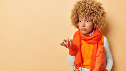 Puzzled confused woman with curly hair shrugs shoulders stares with omg expression dressed in casual clothes carries bag under arm isolated over beige background copy space for your promotion