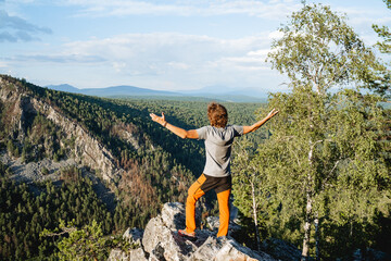 A happy guy stands in the mountains with his hands to the sides, a hipster climbed a stone, a victorious raising of his hands to the sky, a beautiful view of the mountains, a man a view from behind.